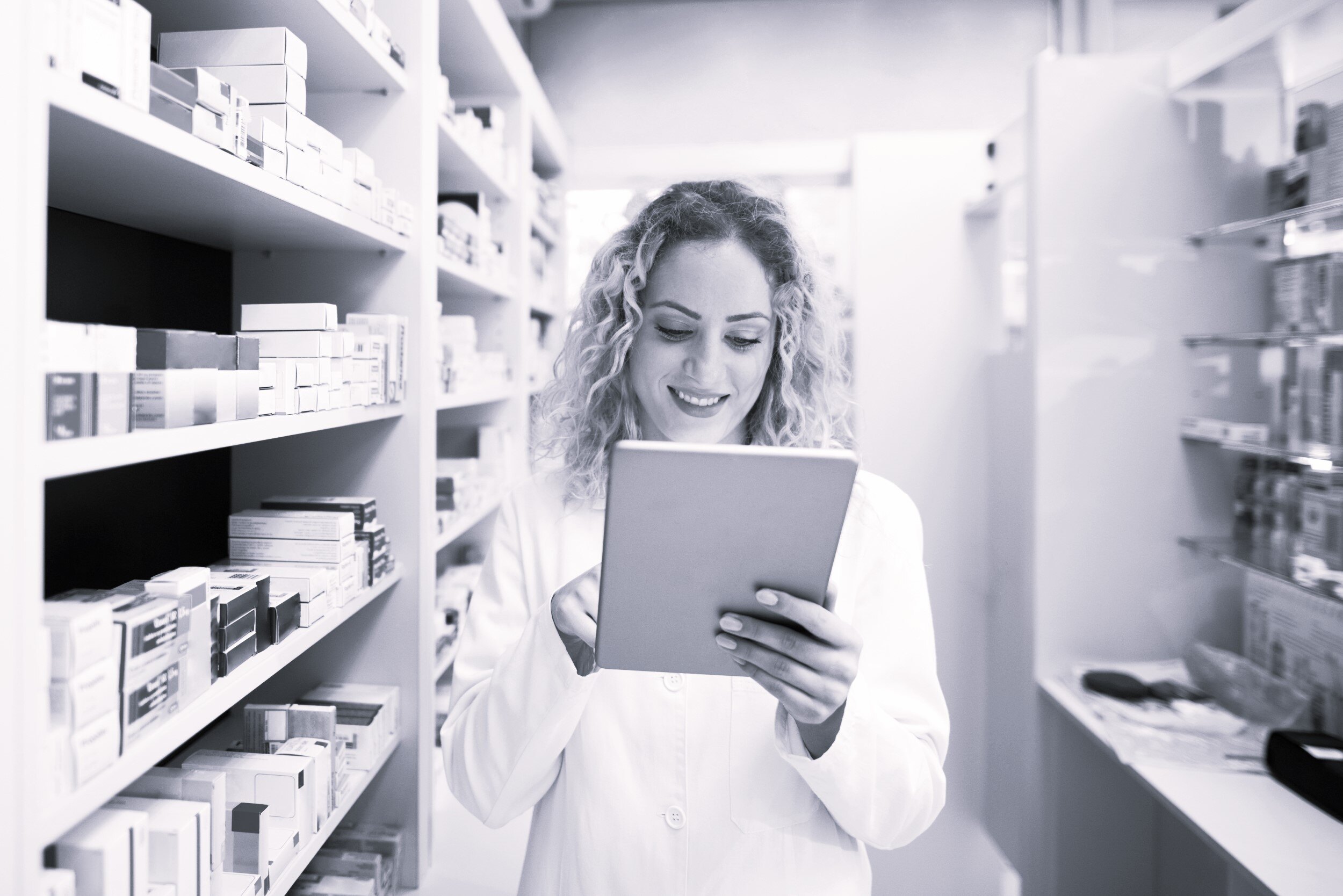 Smiling pharmacist standing between shelves of medicine, using a digital tablet to check pharmaceutical inventory and patient information.